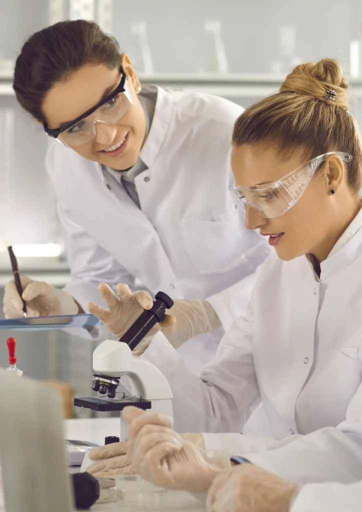 Two scientists, wearing white lab coats and protective eyewear, work closely in a well-lit laboratory setting. One examines a sample under a microscope while the other makes notes. Both exhibit focused concentration on their tasks.