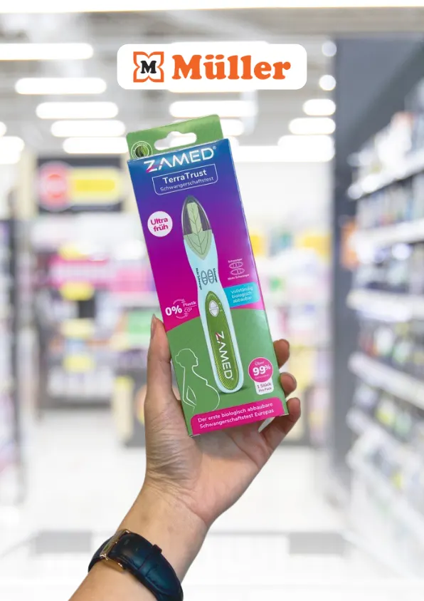 A person's hand holding up a box of ZAMED Terra Trust eco pregnancy test in a store aisle. The box is prominently green and purple with text highlighting the product as 'ultra early,' '0% plastic' and '99% accurate.